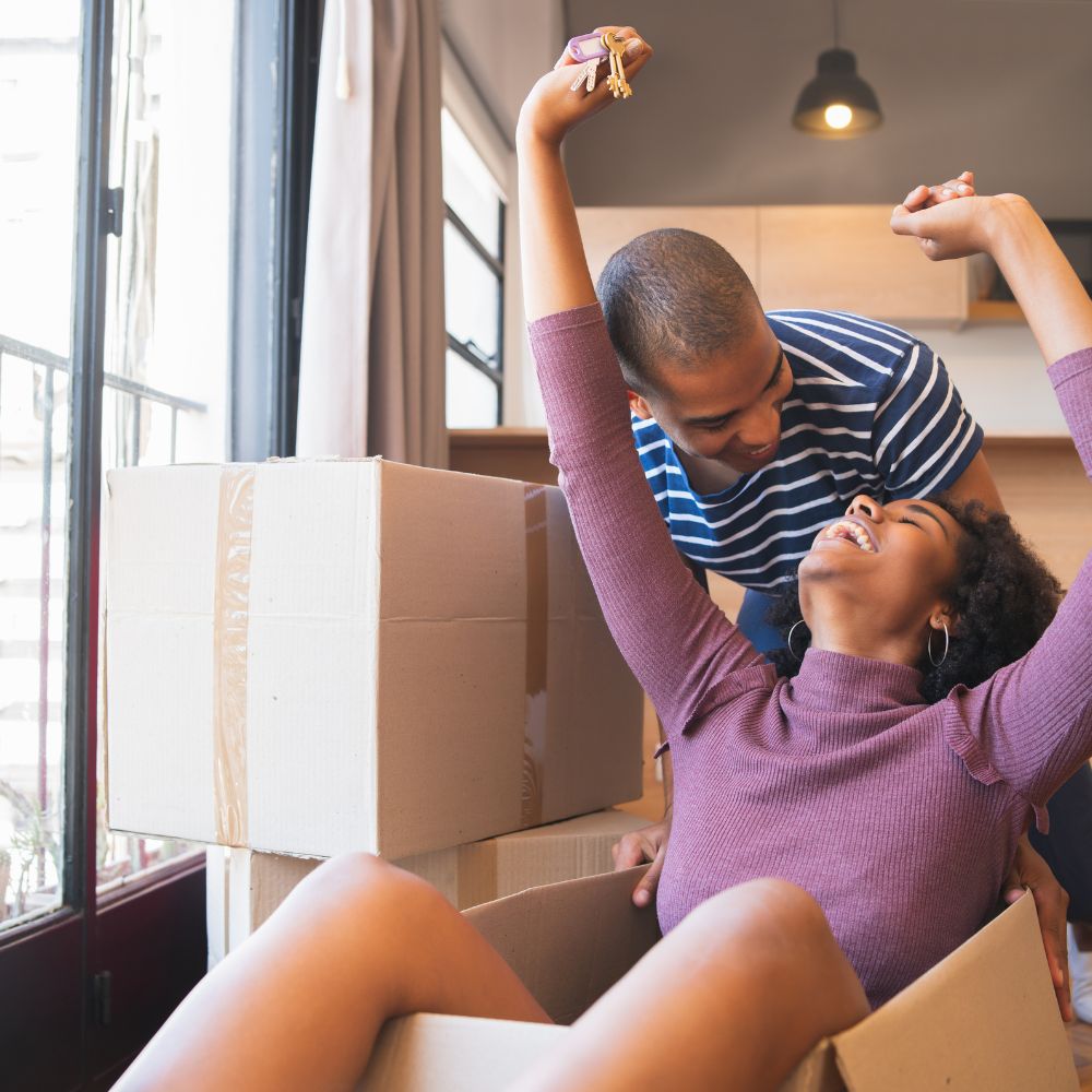 Excited wife sitting in cardboard moving box holding house keys with her loving husband looking down at her