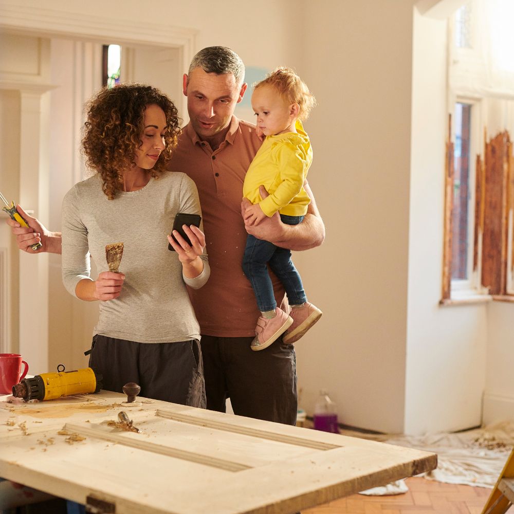 Husband holding small child and paint roller while wife shows him ideas on phone surrounded by home repairs